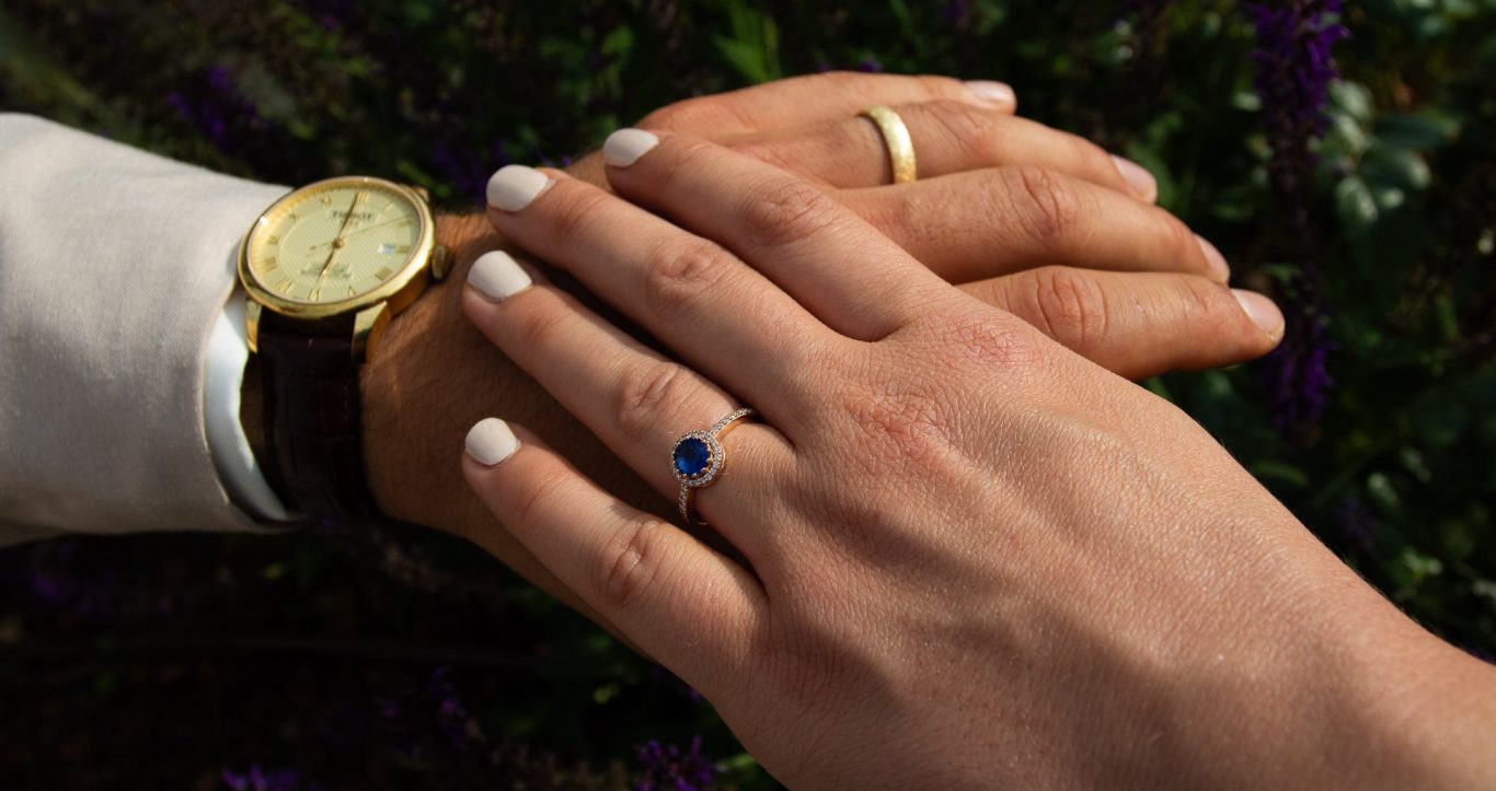 Hände mit Ringen, eine mit blauem Stein, auf einem Hintergrund aus blühenden Pflanzen.