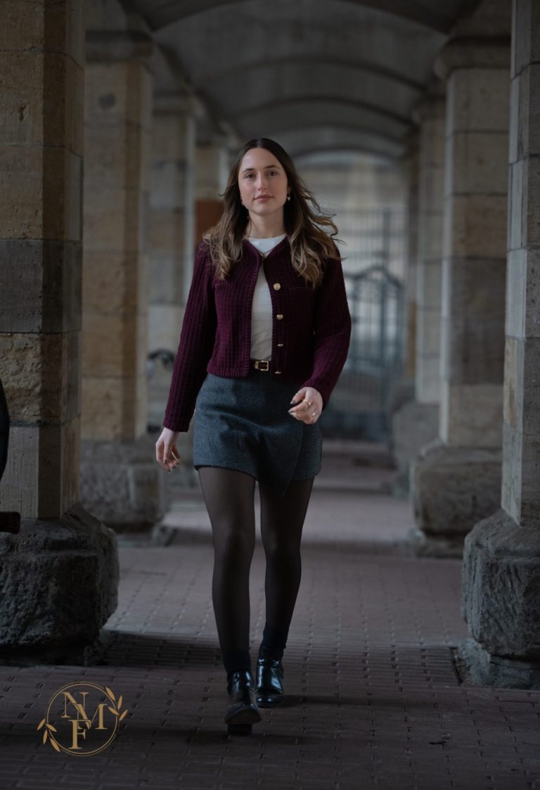Eine Frau in einer burgunderfarbenen Jacke und Rock geht durch einen architektonischen Gang.