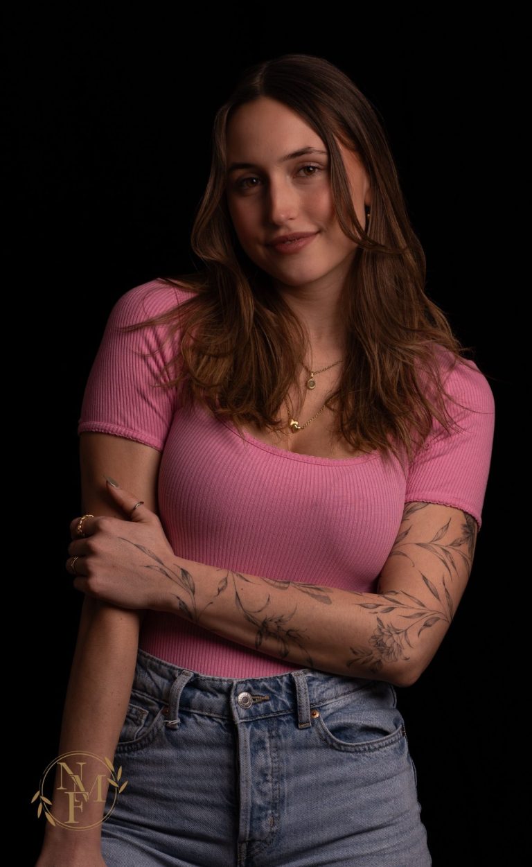 Junge Frau mit langen Haaren, im rosa Shirt und blauer Jeans, mit tätowierten Armen.
