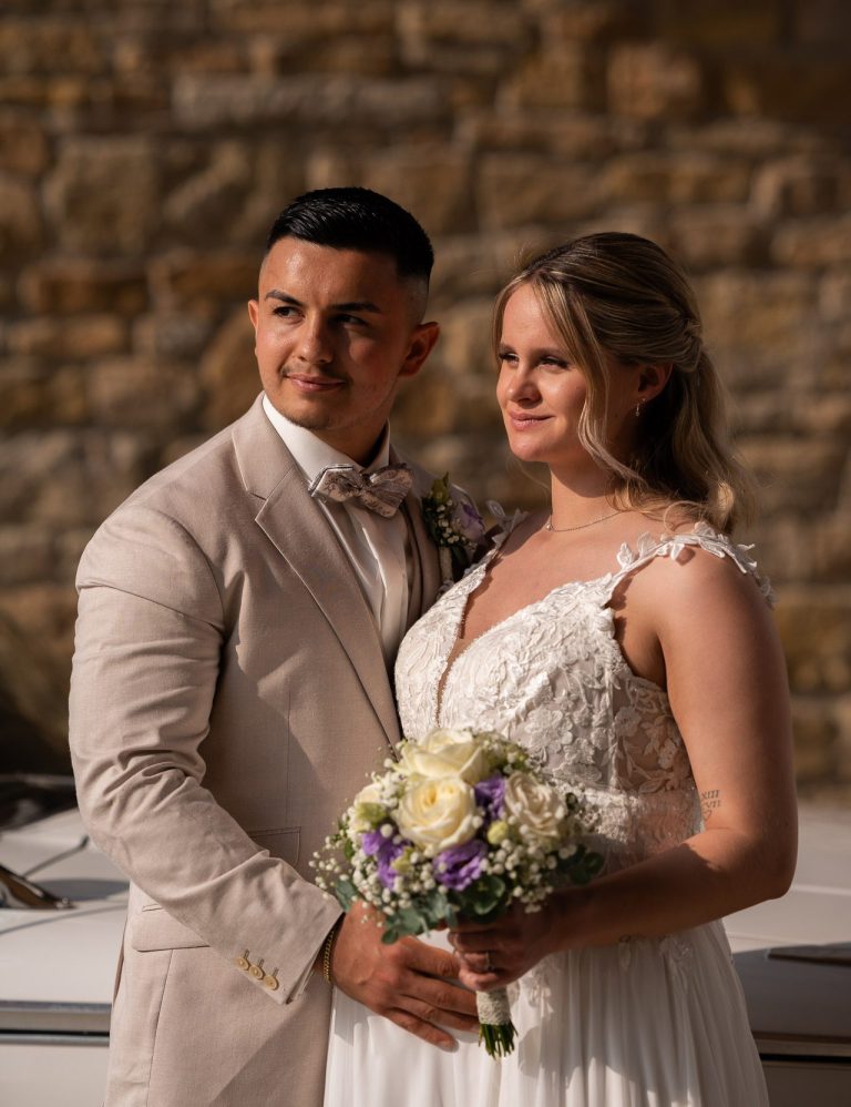 Ein Brautpaar in Hochzeitskleidung steht lächelnd mit einem Blumenstrauß.