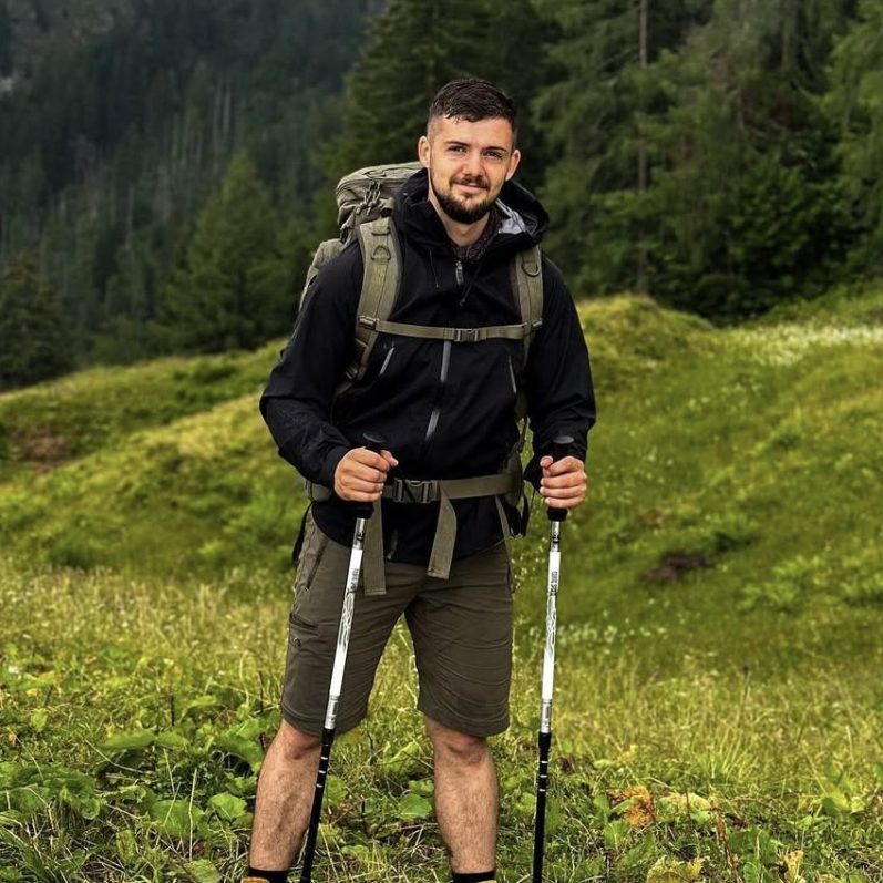 Mann mit Wanderausrüstung steht in einer grünen Landschaft.