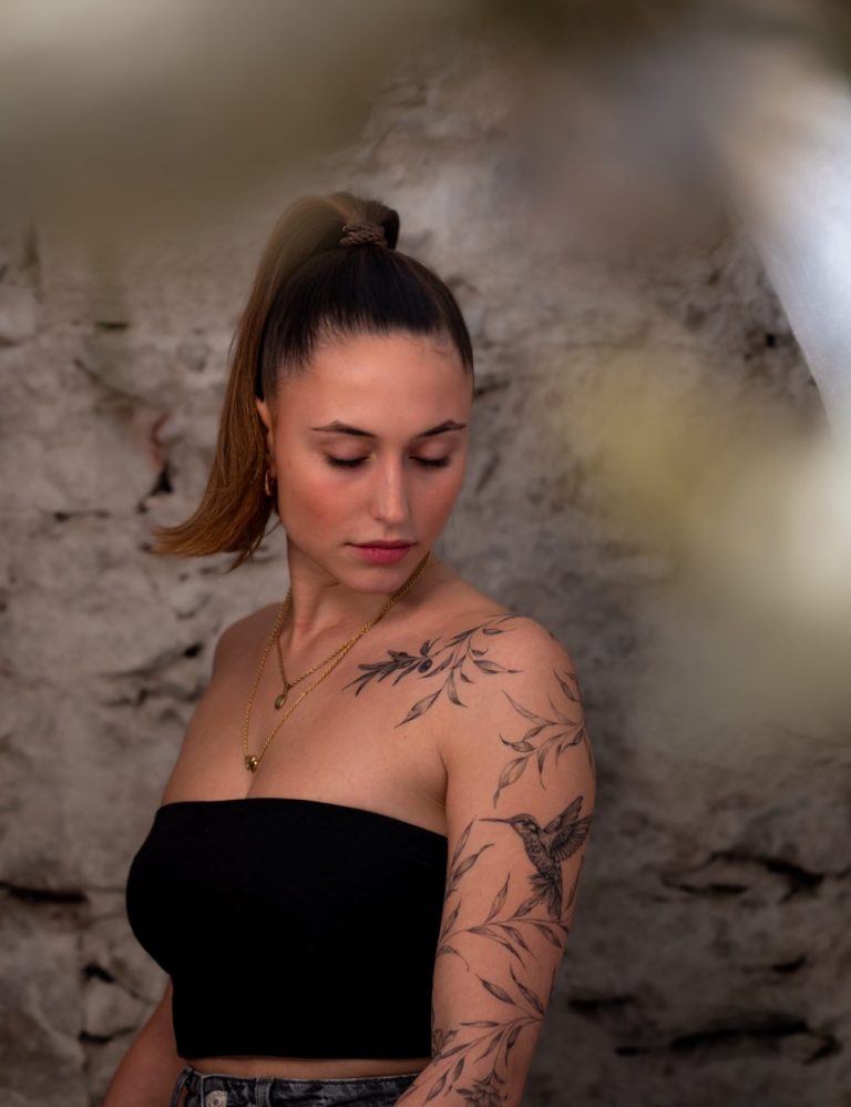 Junge Frau mit einem schwarzen Oberteil und einem detaillierten Arm-Tattoo, schaut nach unten.