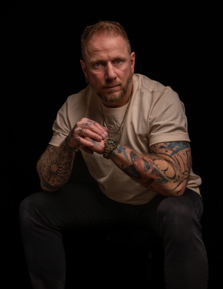 Mann mit Tattoos sitzt in dunkler Umgebung, ernst und nachdenklich blickend.