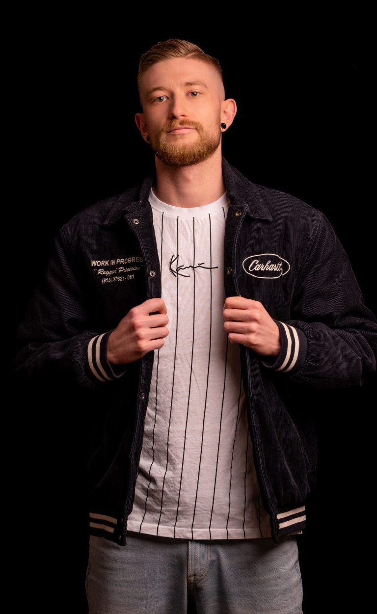 Mann in weißem T-Shirt und schwarzer Jacke mit gestreiften Ärmeln, lächelt leicht.