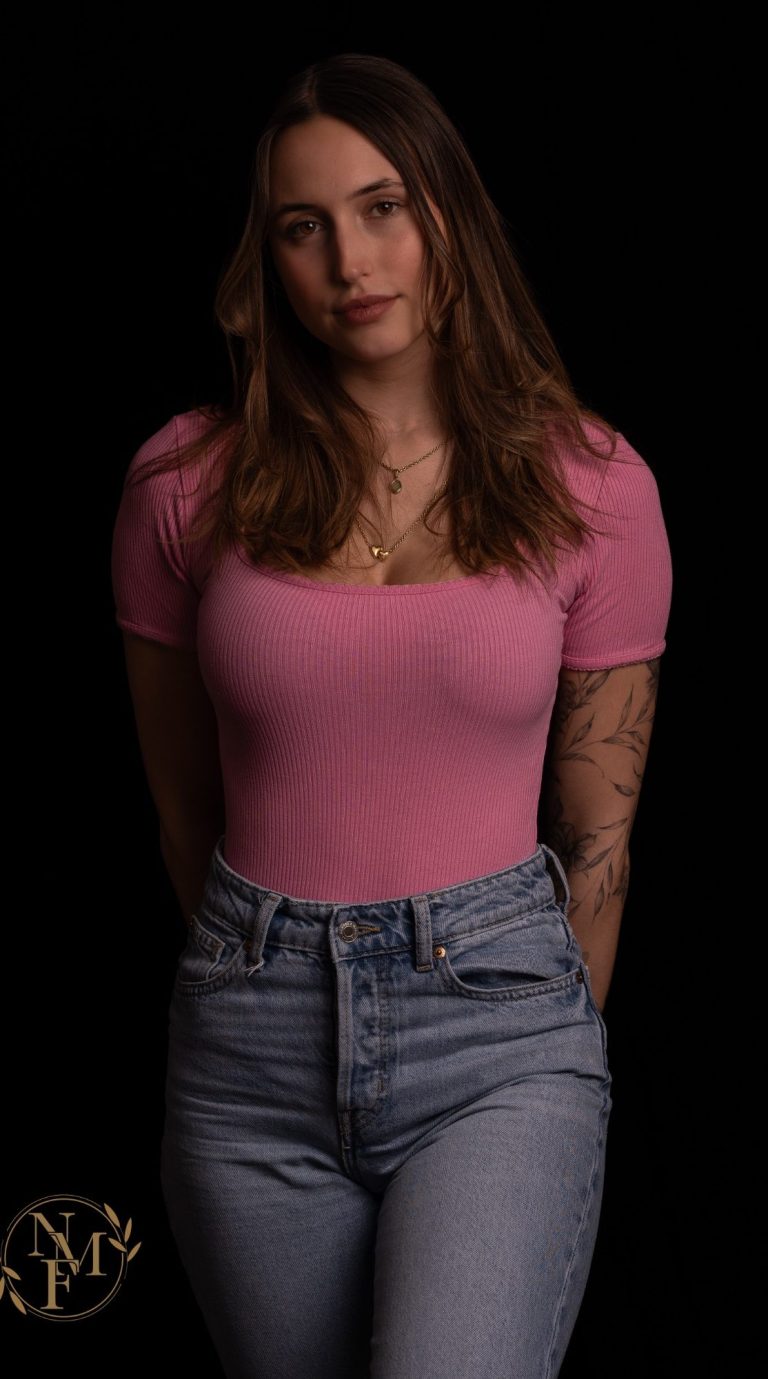Frau mit langen braunen Haaren in rosa Oberteil und Jeans, vor schwarzem Hintergrund.