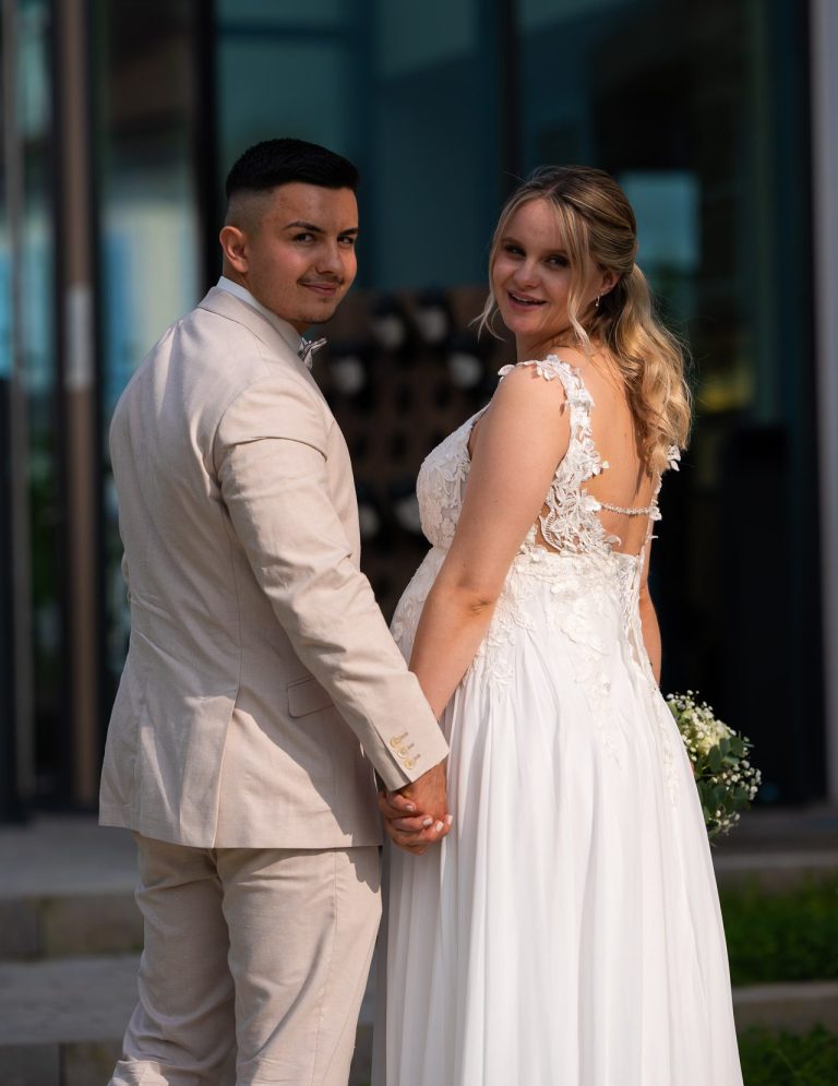 Brautpaar hält Händchen vor modernem Gebäude, beide lächeln in Hochzeitskleidung.