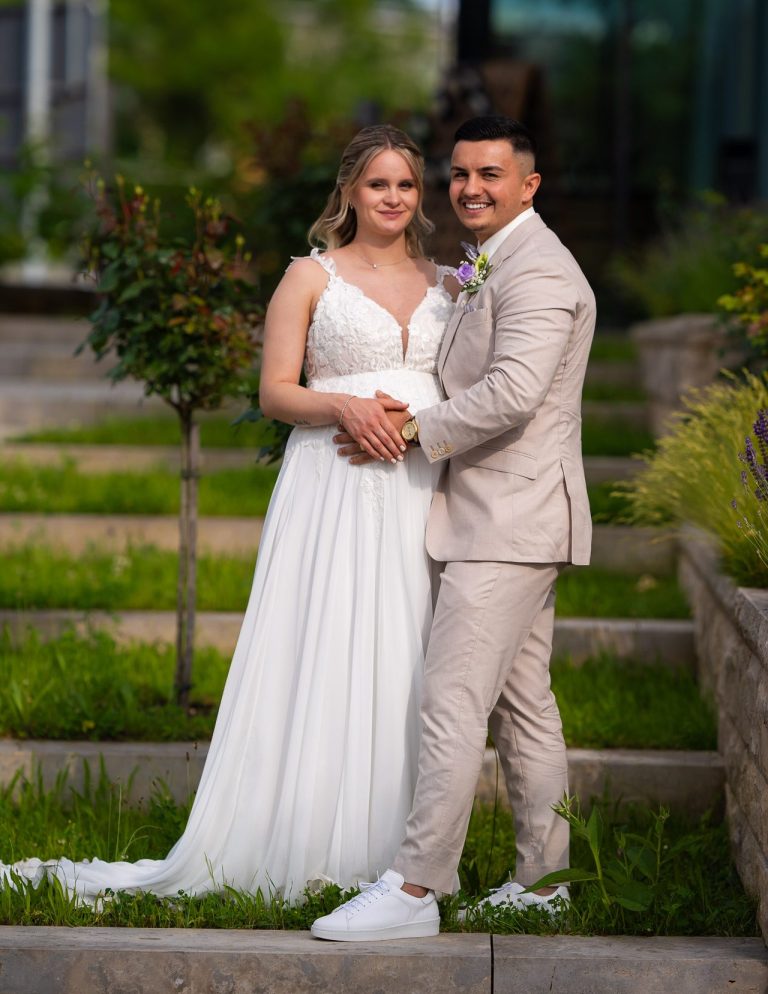 Hochzeitscouple in elegant Kleidung steht lächelnd vor grüner Gartenkulisse.