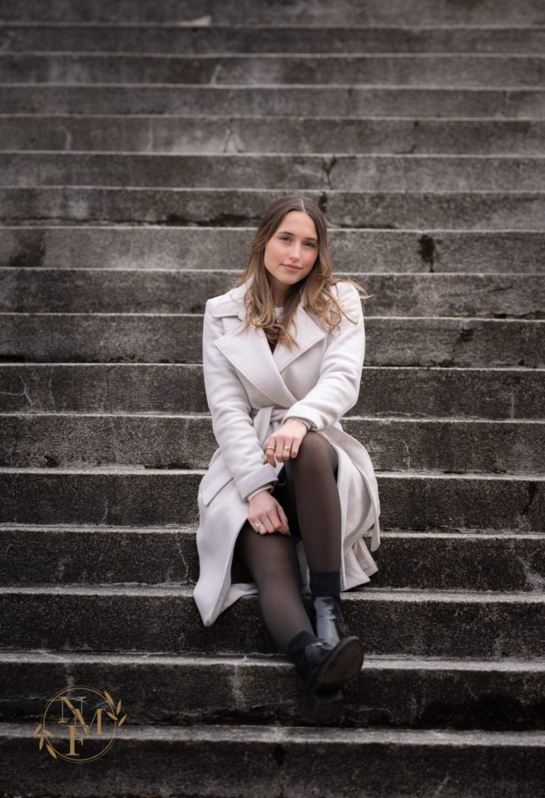 Junge Frau in einem grauen Mantel sitzt auf einer steinernen Treppe.