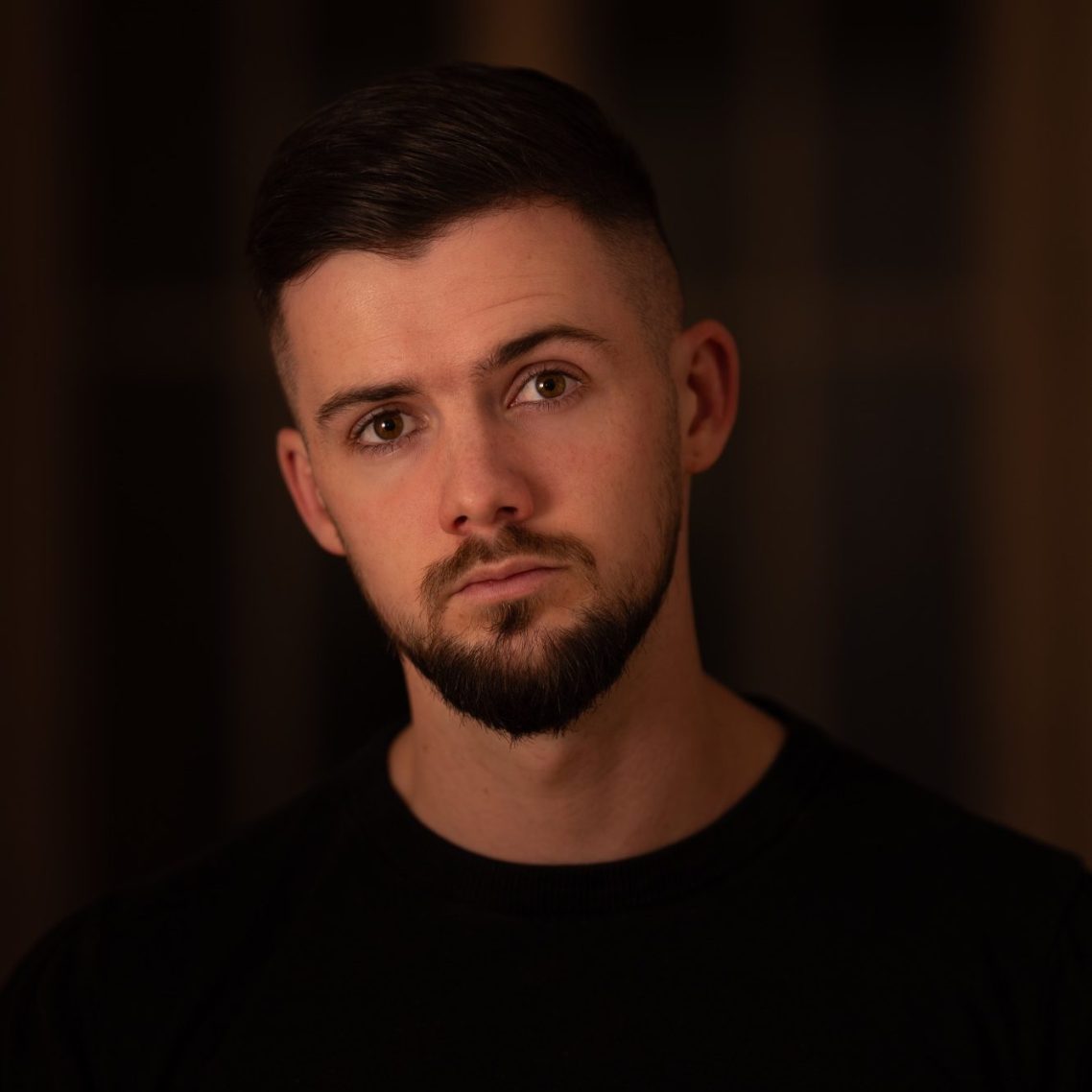 Junger Mann mit kurzem Haar und Bart, schaut ernst in die Kamera.