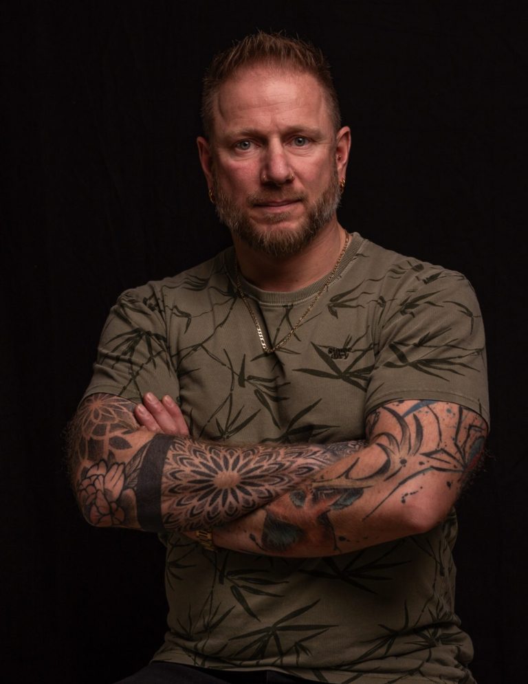 Mann mit tatowierten Armen, in einem schwarzen Shirt, mit ernstem Blick.